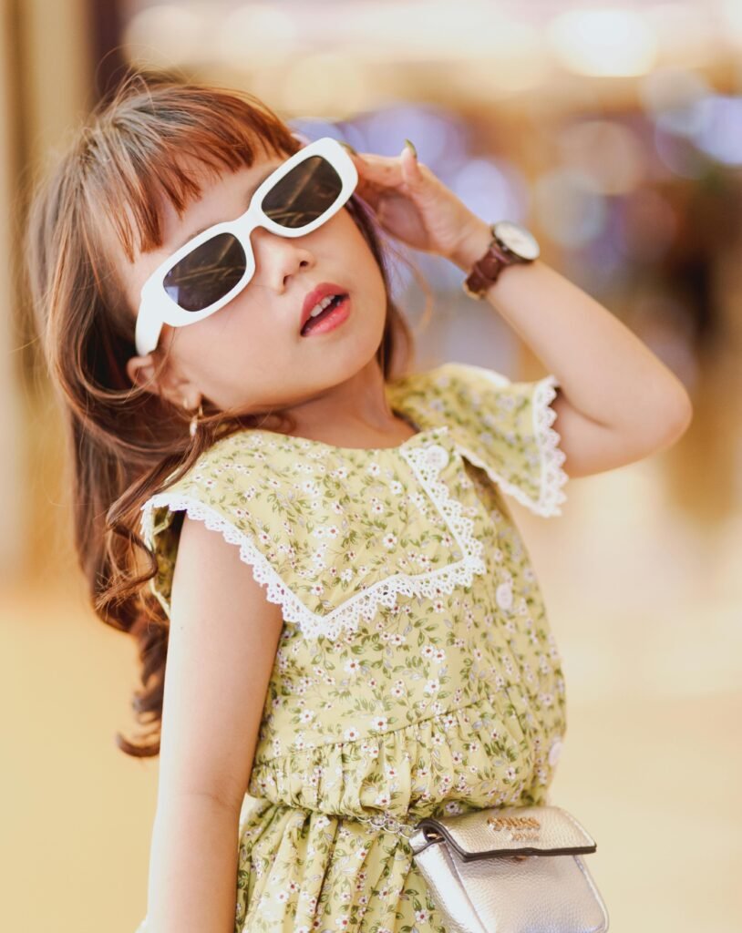 Home Adorable young girl stylishly posing indoors wearing sunglasses and a floral dress.